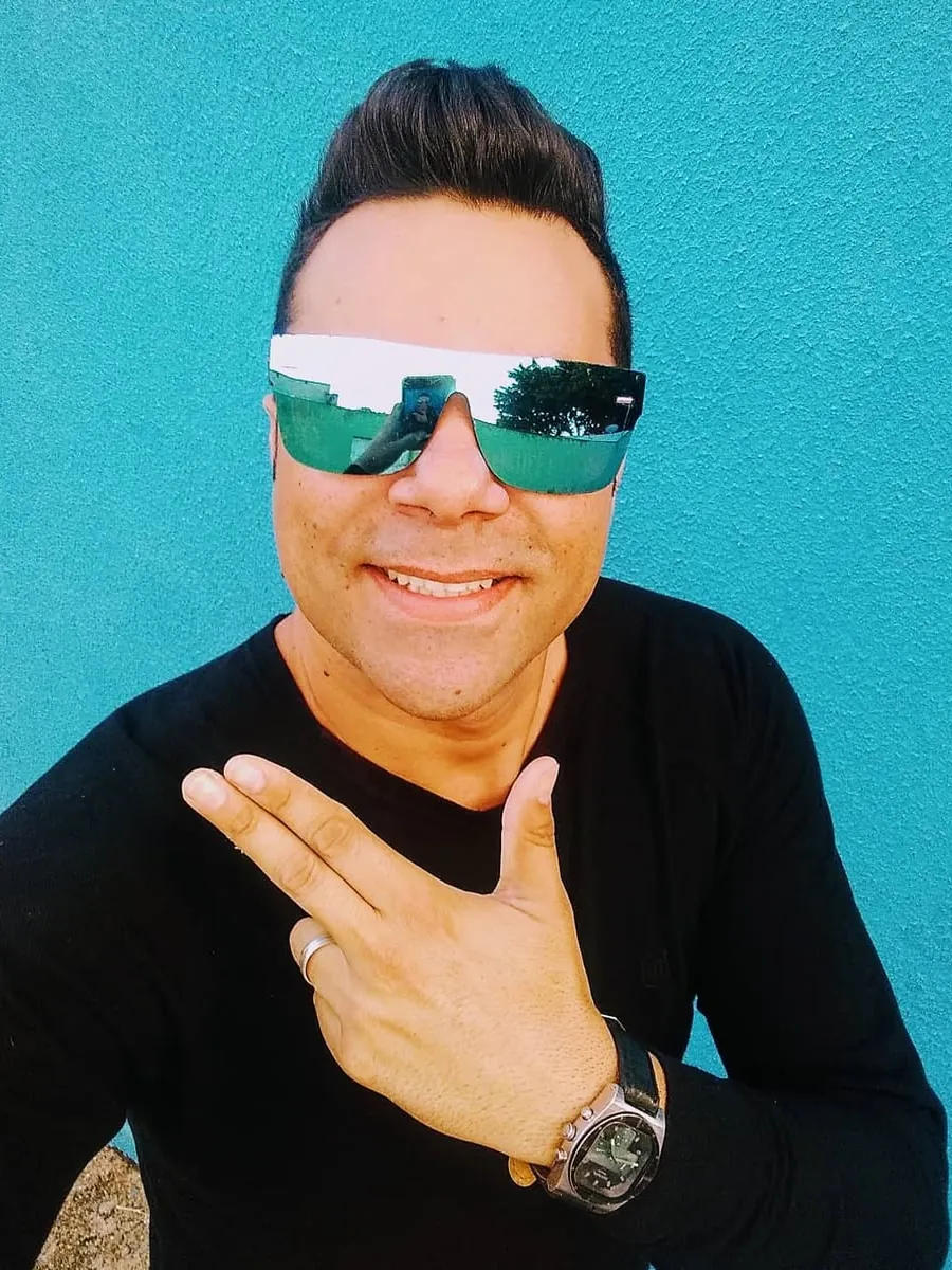 Luciano Lins sorrindo, de óculos espelhados, em frente a uma parede azul